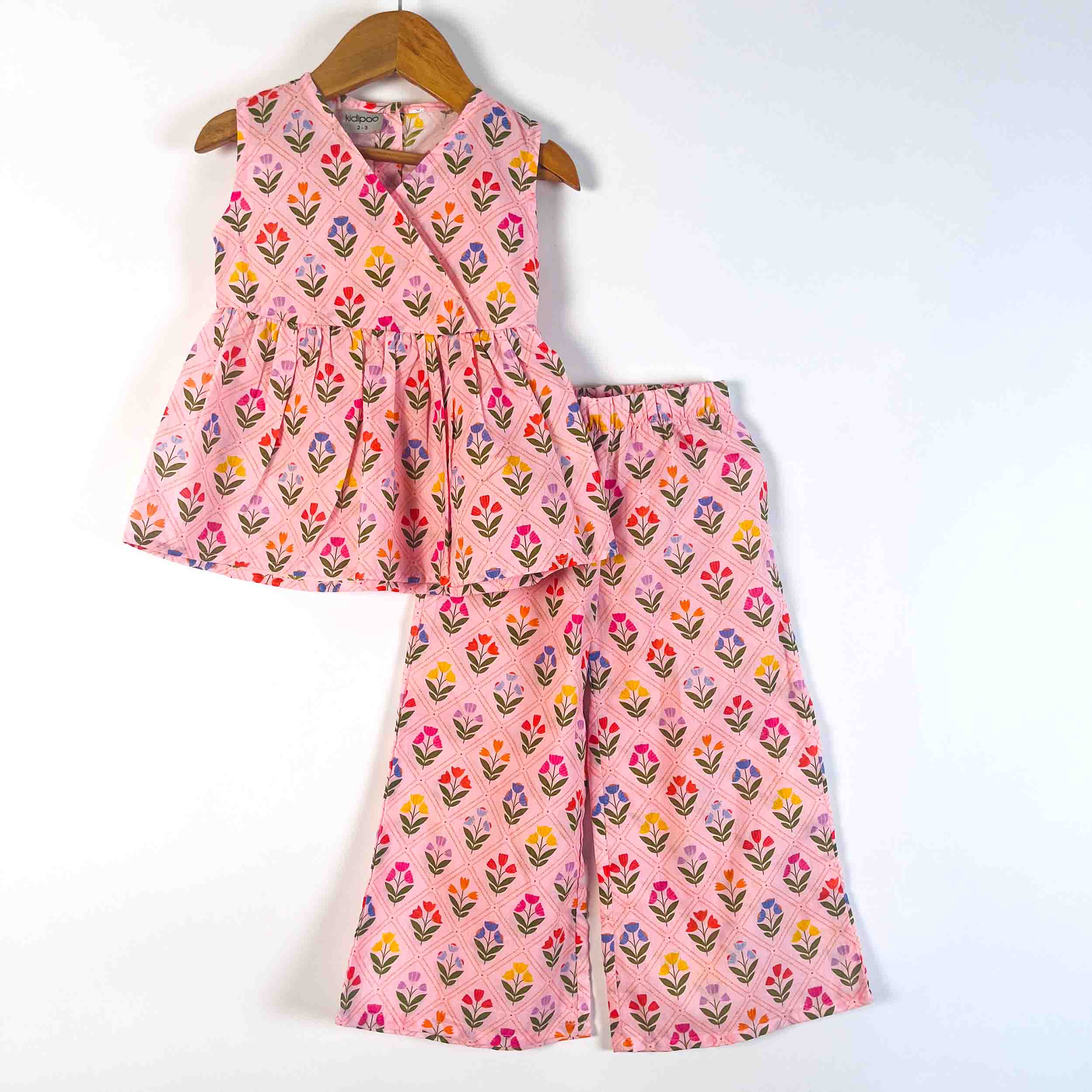 A two-piece girls' outfit set consisting of a sleeveless wrap-style top and matching wide-leg pants, both in pink with a colorful floral block print, hanging on a wooden hanger against a plain white background.