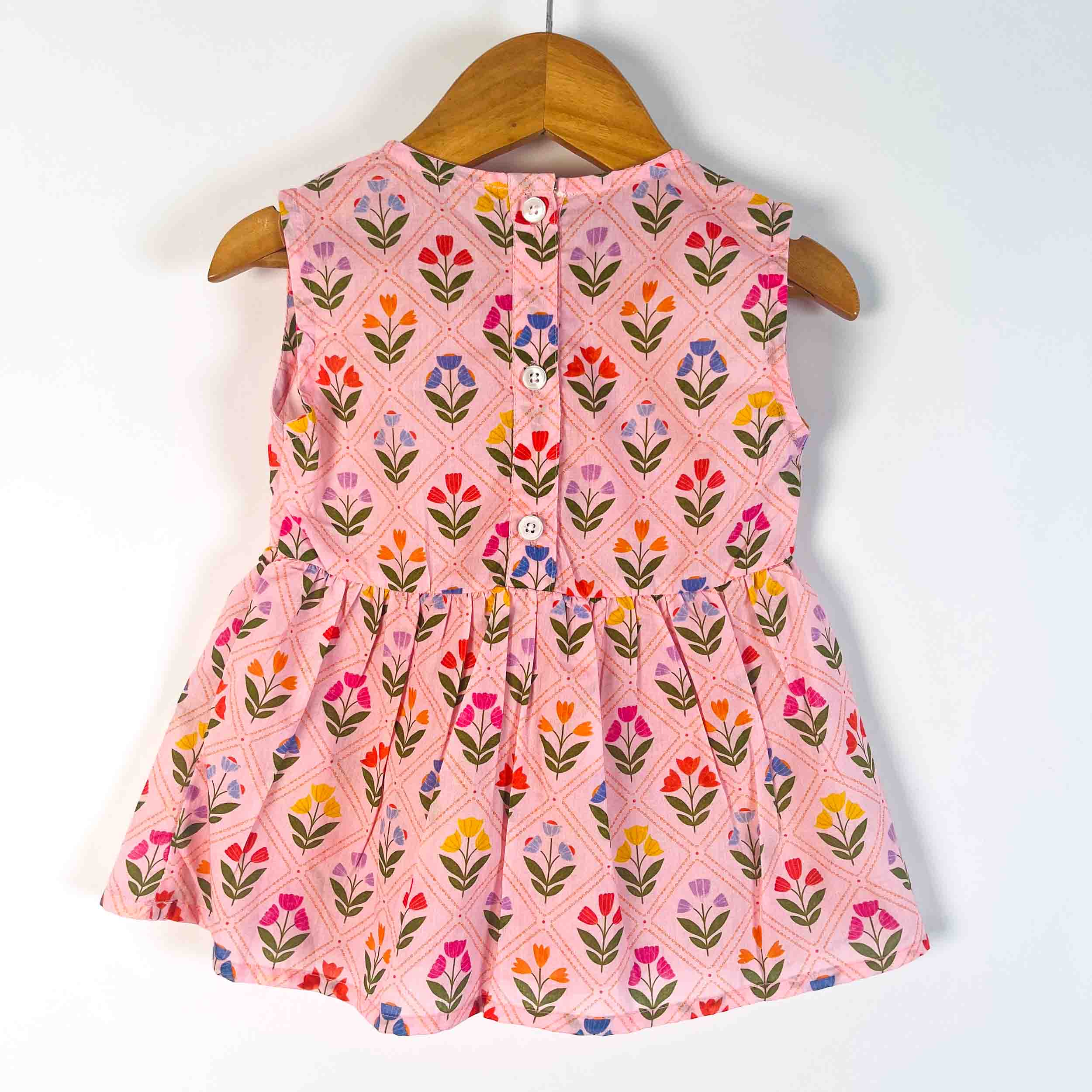 A sleeveless pink children's dress with a colorful floral and geometric pattern, hanging on a wooden hanger against a plain white background.
