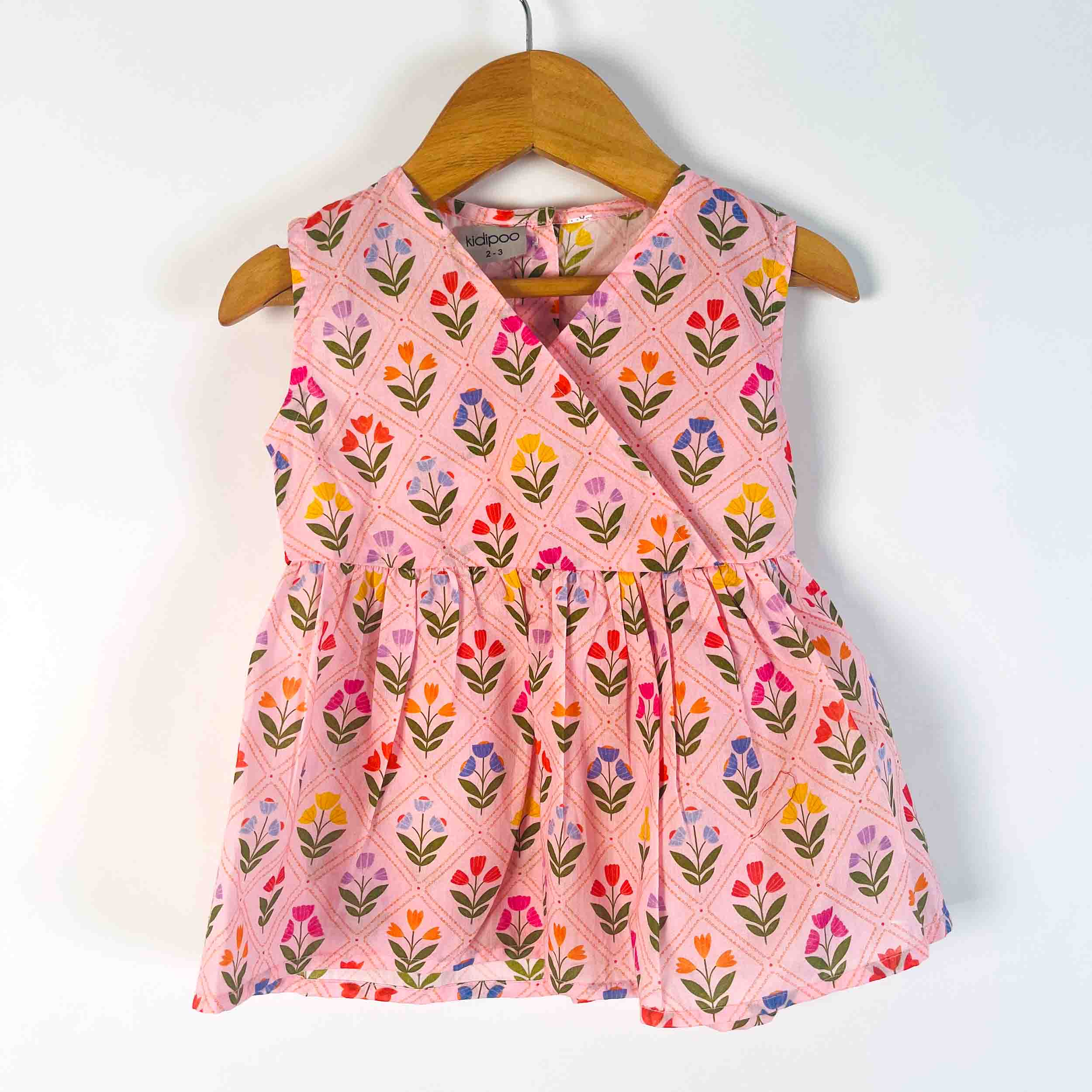 A sleeveless pink wrap-style dress with a colorful floral and geometric print, hanging on a wooden hanger against a plain white background.