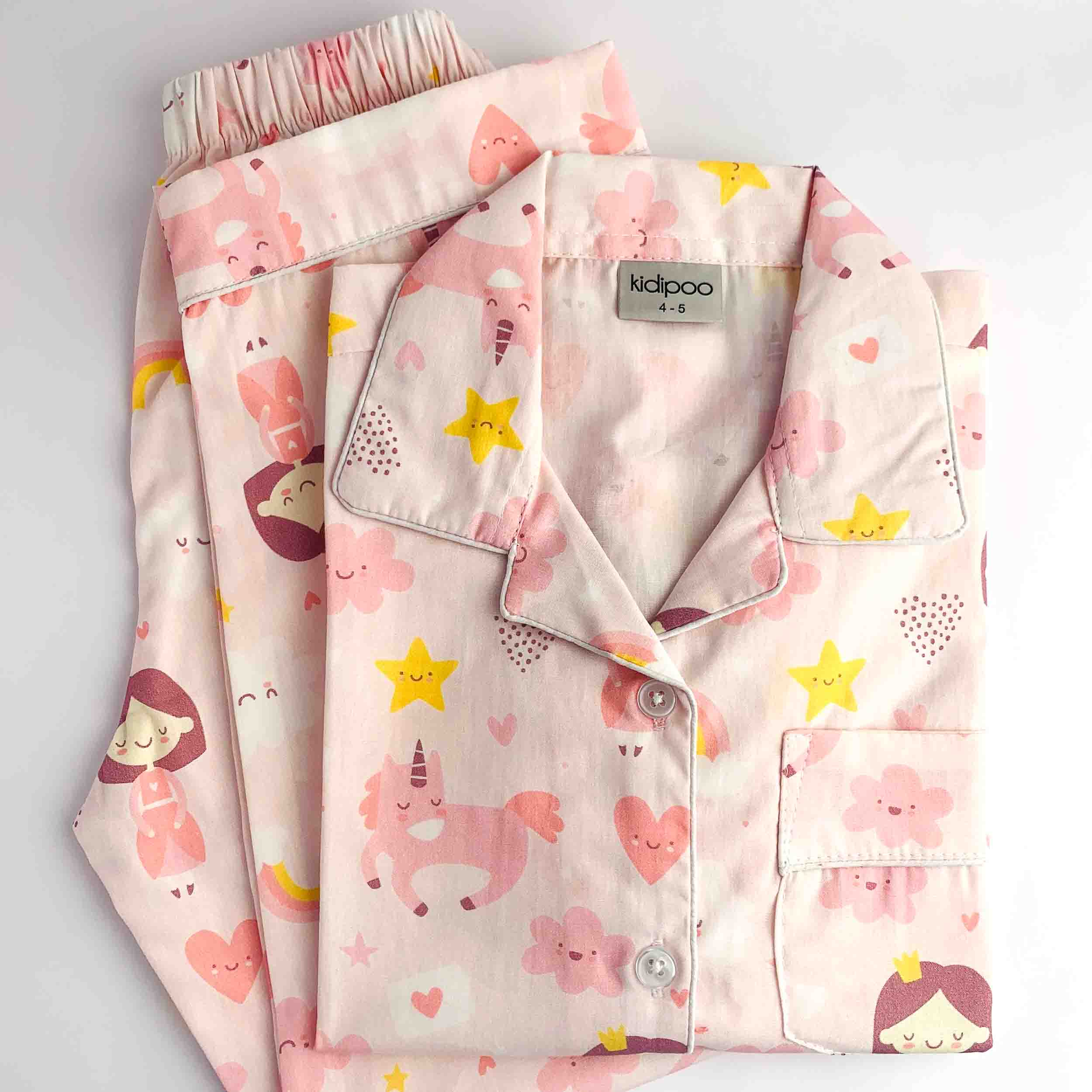 A folded children's pajama set with matching shorts, featuring a light pink base with a playful print of unicorns, hearts, stars, and cartoon girls. The brand label 'kidpoo' is visible inside the collar.