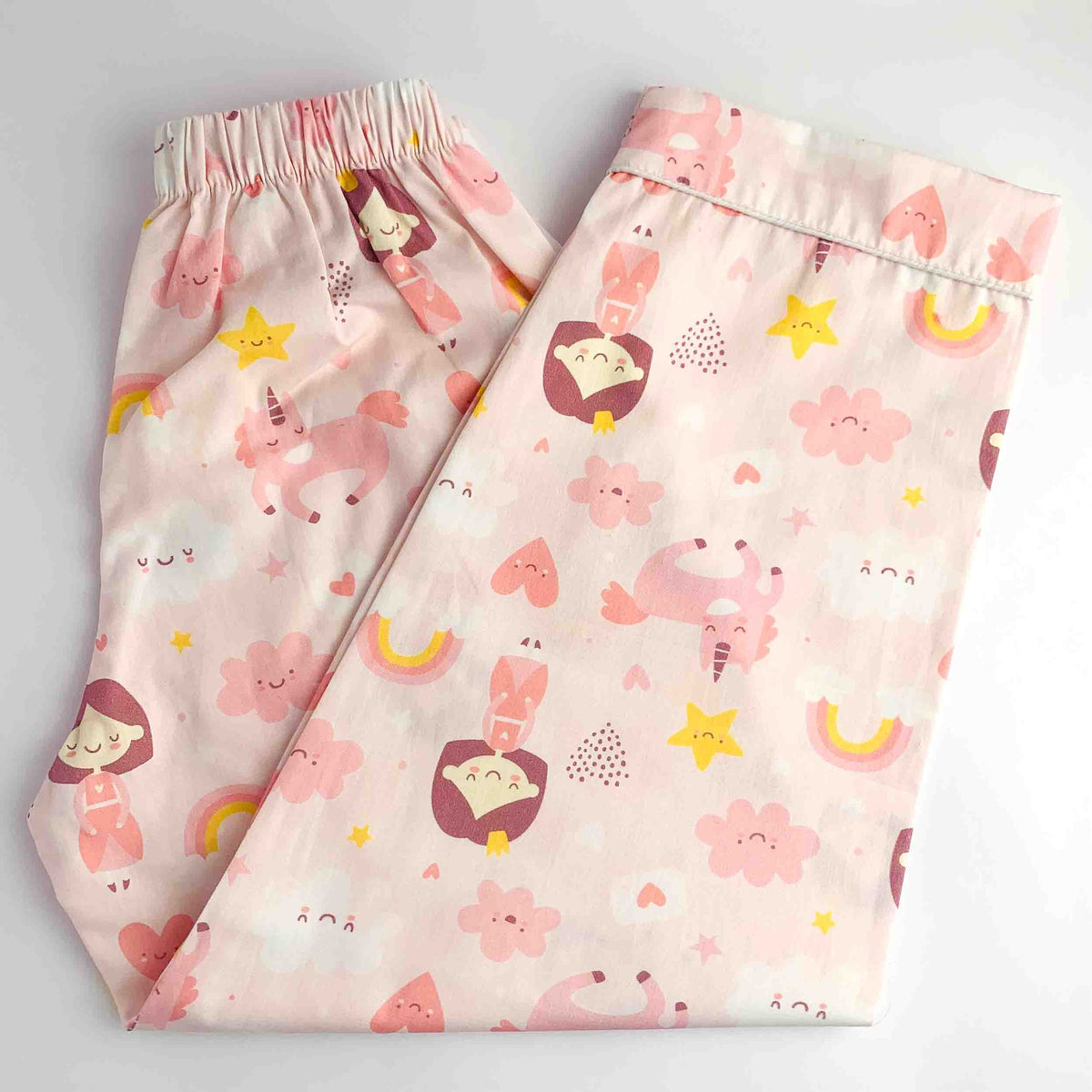 A pair of light pink pajama pants with a whimsical cartoon unicorn and rainbow print, folded neatly on a white background.
