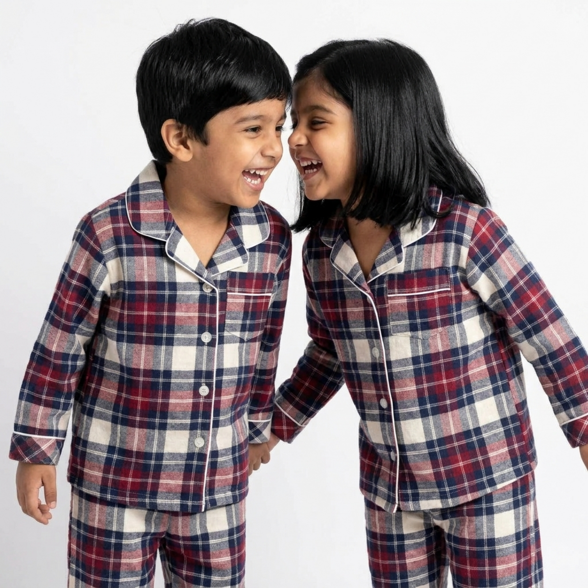 Two children wearing matching long-sleeve plaid pajamas in red, navy, and white, standing close together against a plain light background.