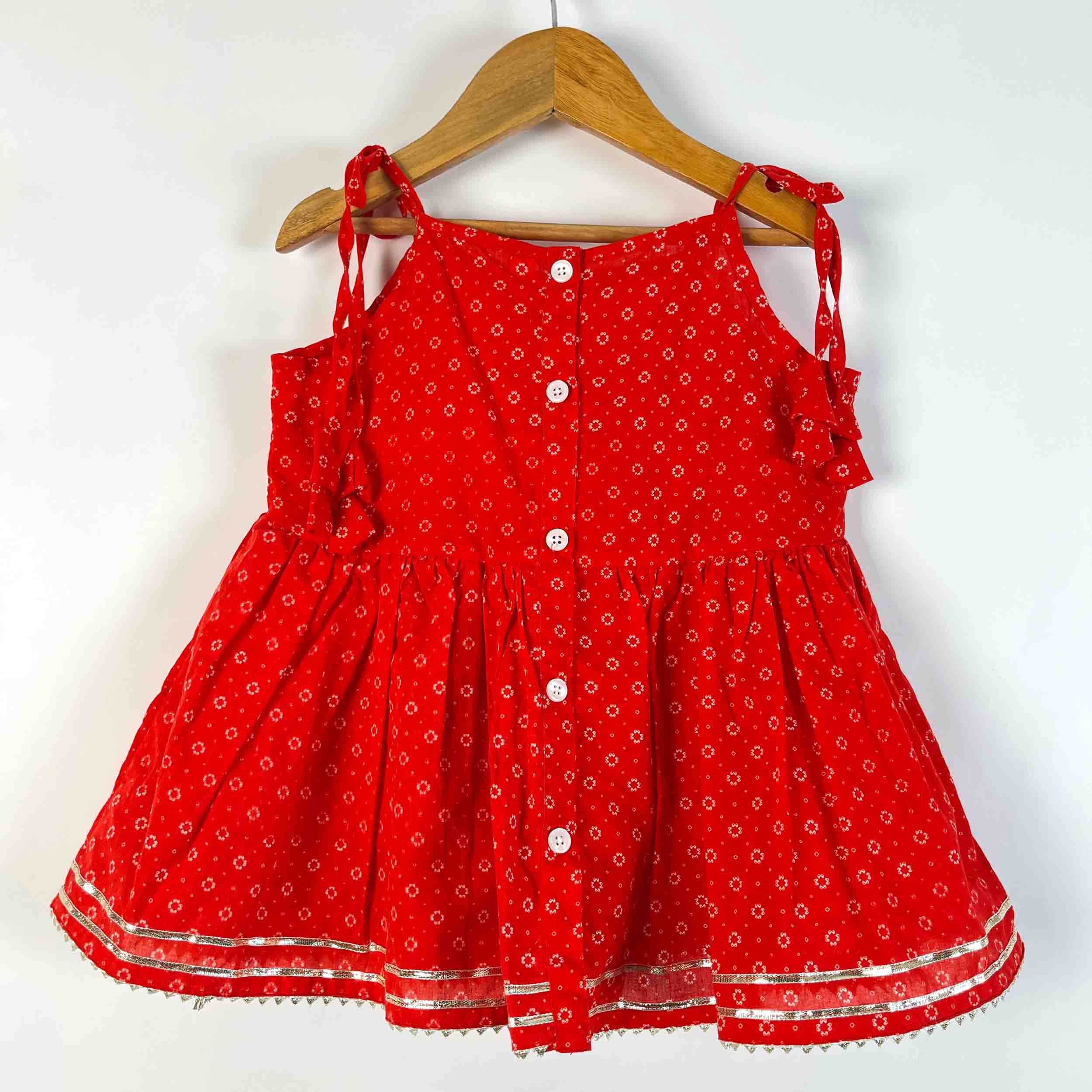A red sleeveless baby dress with a small white floral print, featuring a button-down front, ruffled shoulder straps, and a flared skirt, hanging on a wooden hanger against a plain white background.