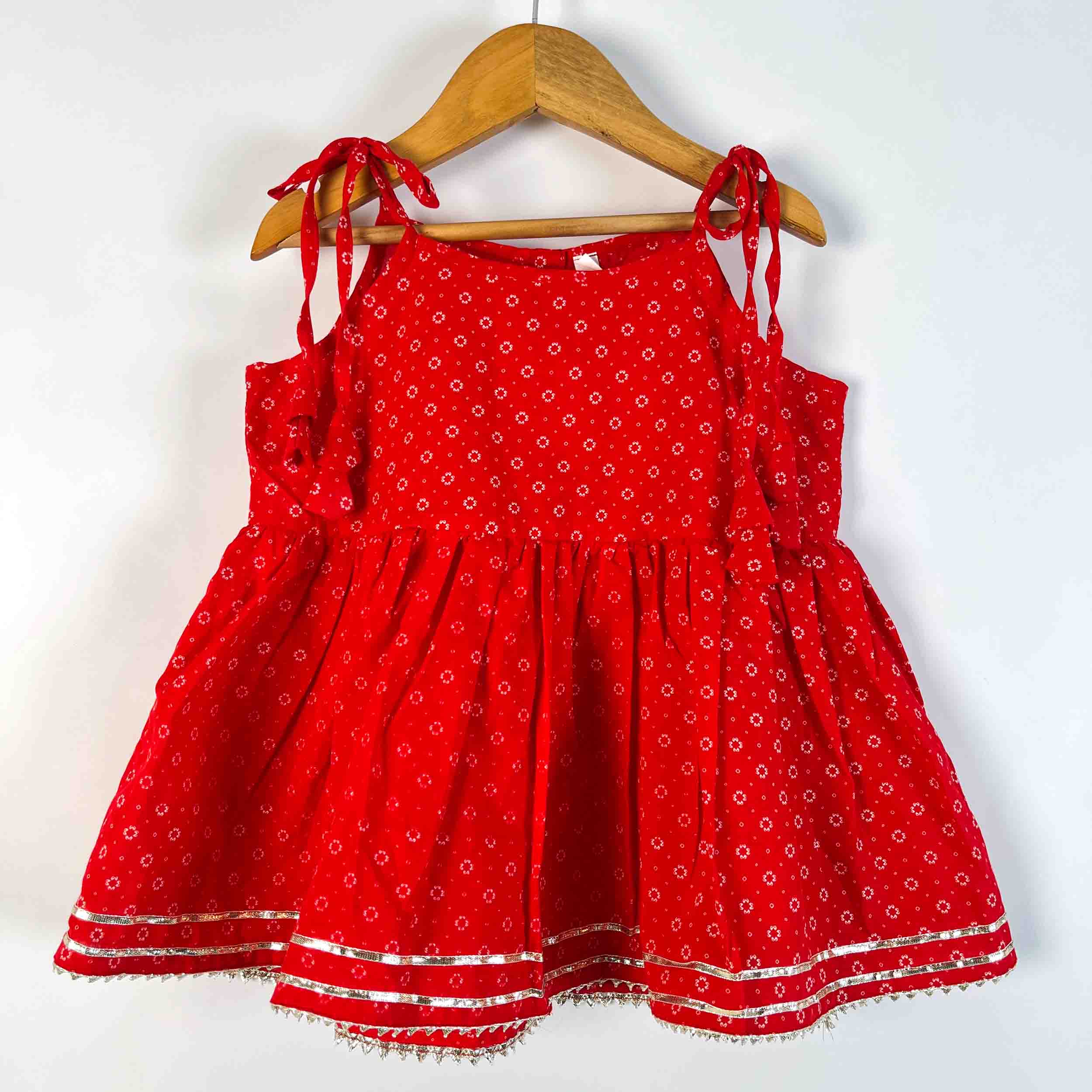 A red sleeveless children's dress with a small white floral print, tie shoulder straps, and a flared skirt, hanging on a wooden hanger against a plain white background.