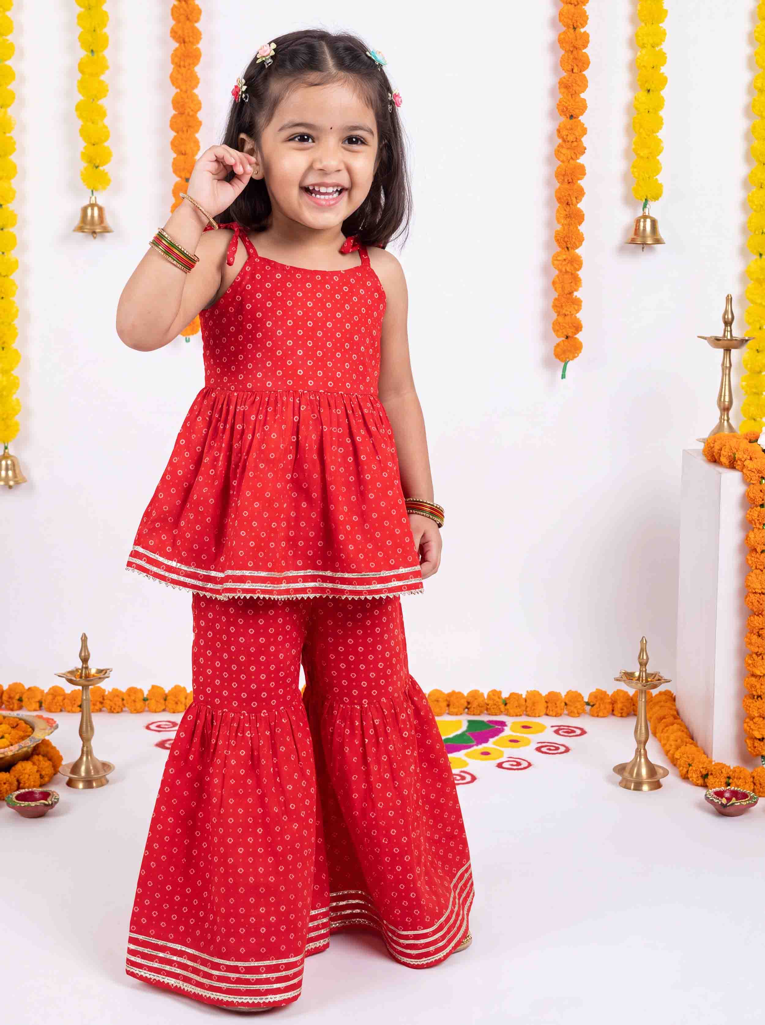 A young girl smiling and posing in a red sleeveless polka dot two-piece outfit with matching flared pants, decorated with marigold garlands and brass lamps.
