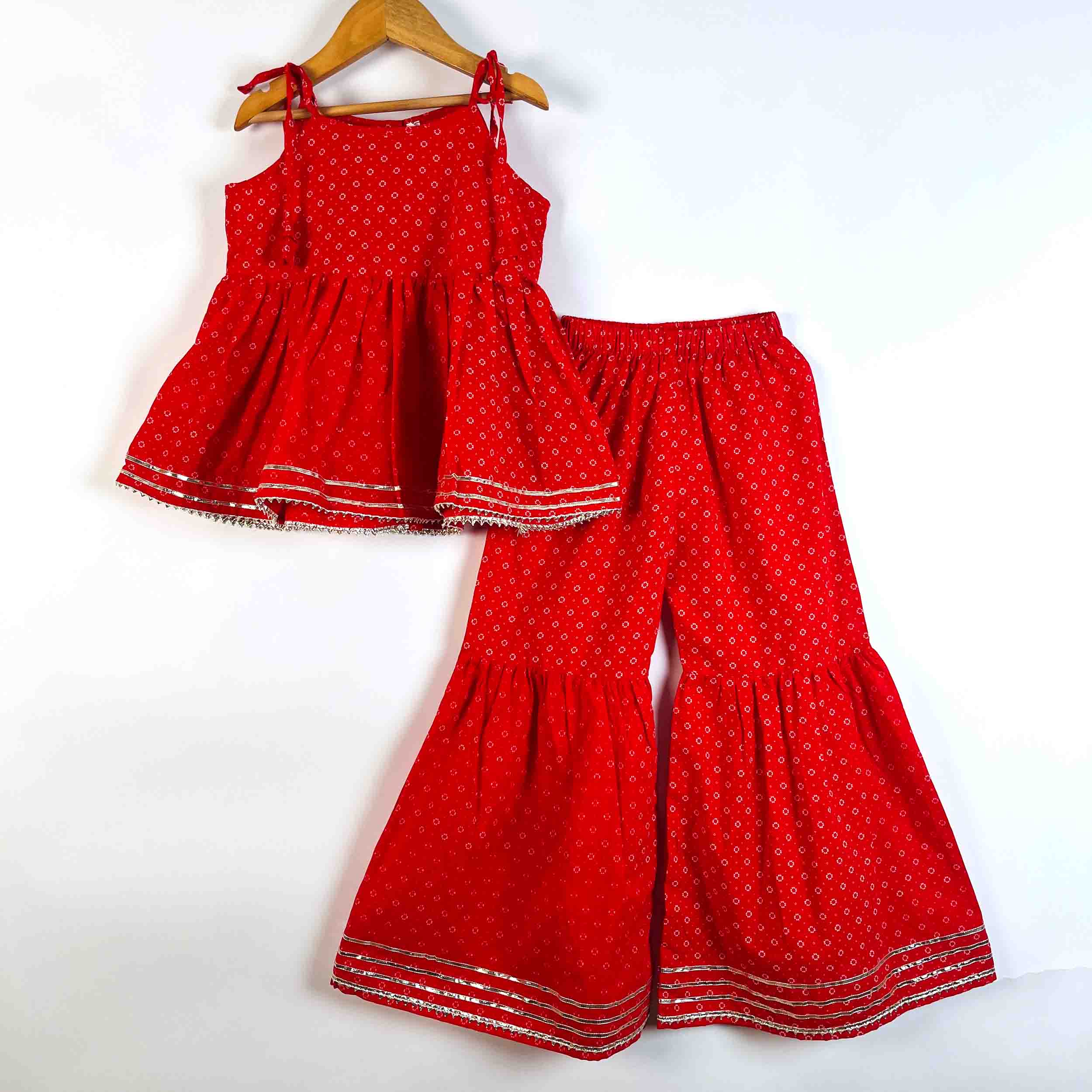 A two-piece children's outfit set consisting of a sleeveless red top with thin shoulder ties and matching wide-leg flared pants, both featuring a small white geometric pattern and silver trim, displayed on a wooden hanger against a white background.