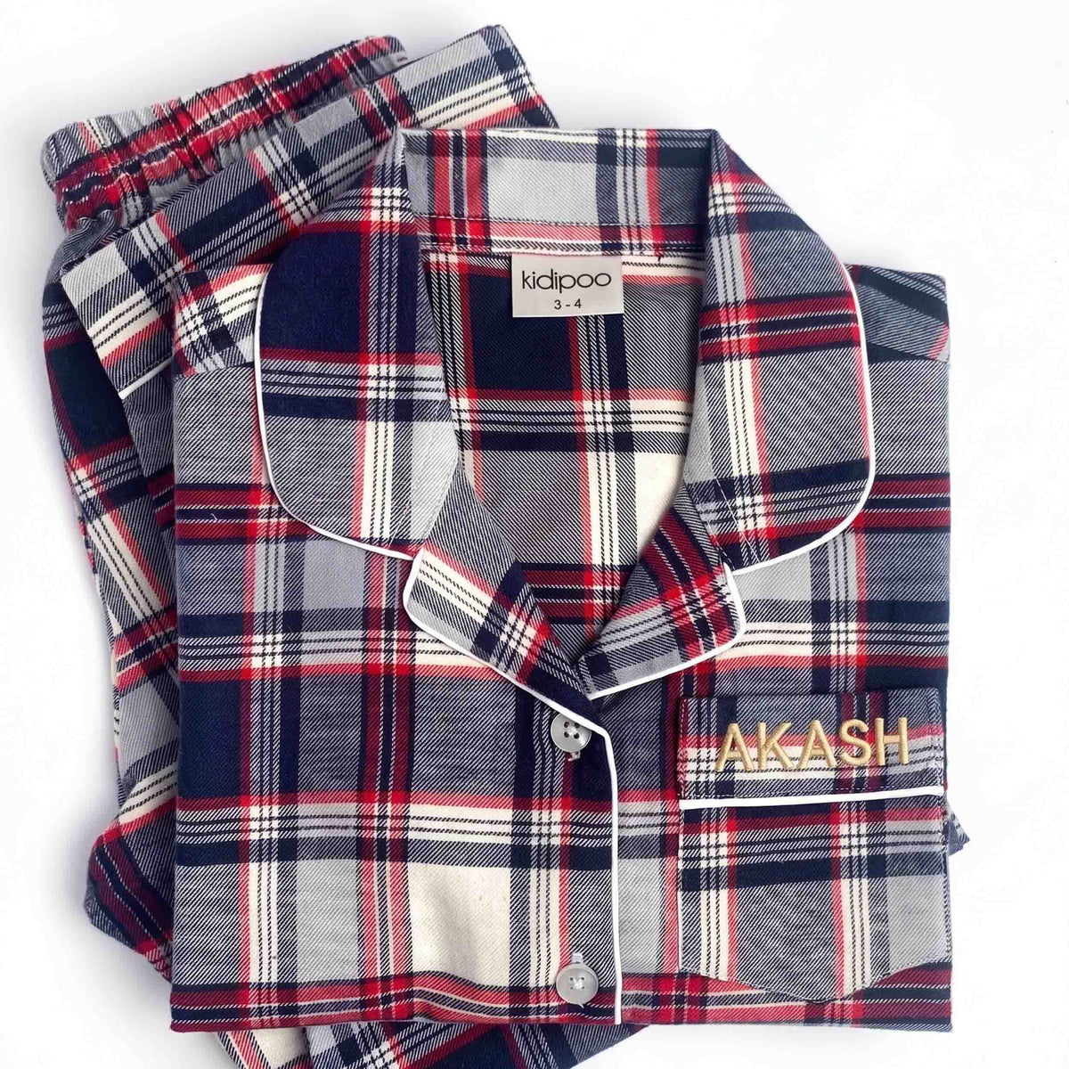 A folded plaid pajama set on a white background, featuring a long-sleeve button-up shirt with a collar and matching pants. The pajamas have a gray, navy, red, and white plaid pattern with white piping.