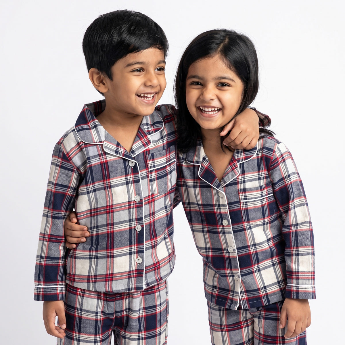 Two children wearing matching plaid pajama sets with navy, red, and white check patterns, standing together against a light gray studio background.