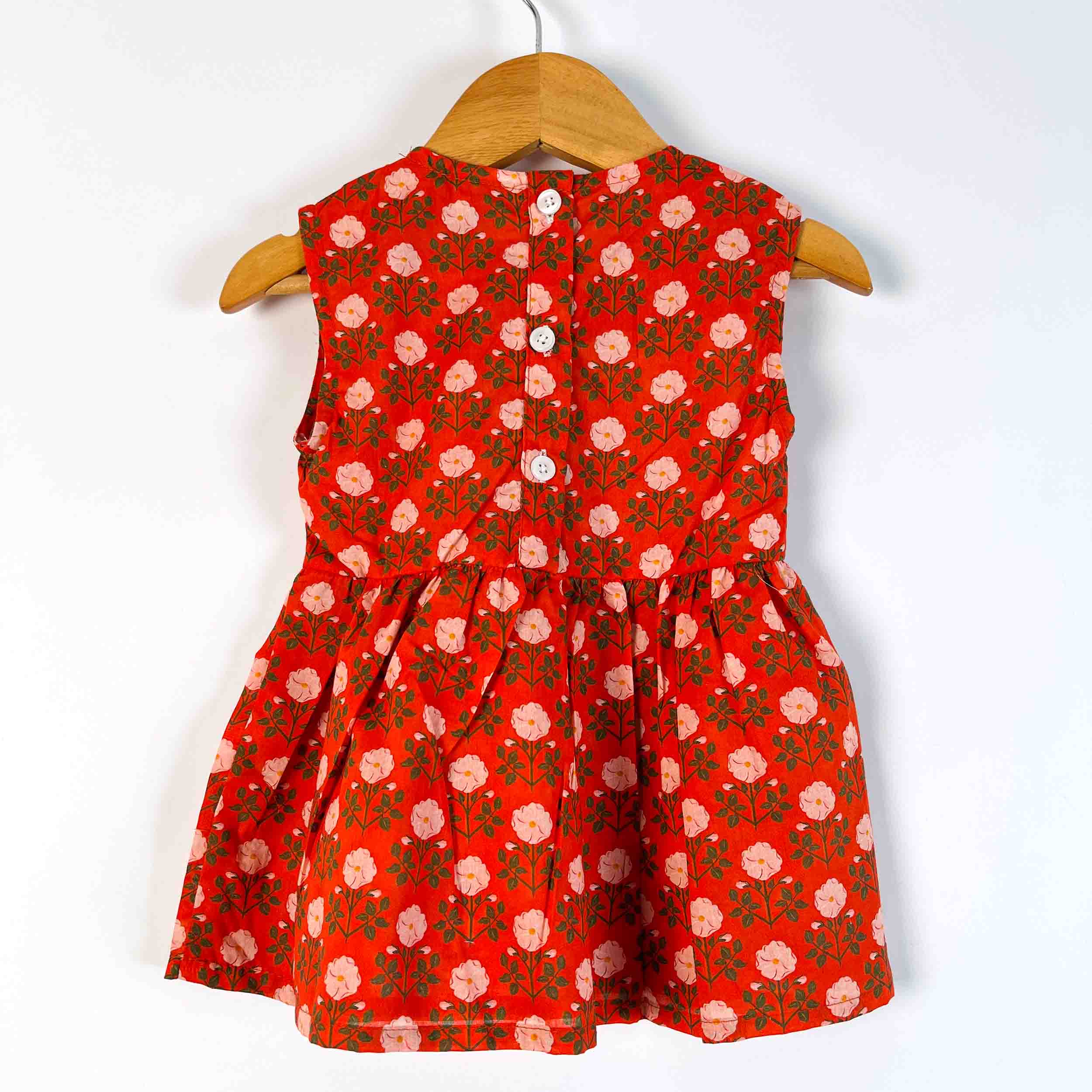 A sleeveless red children's dress with a pink and green floral pattern, hanging on a wooden hanger against a plain white background.