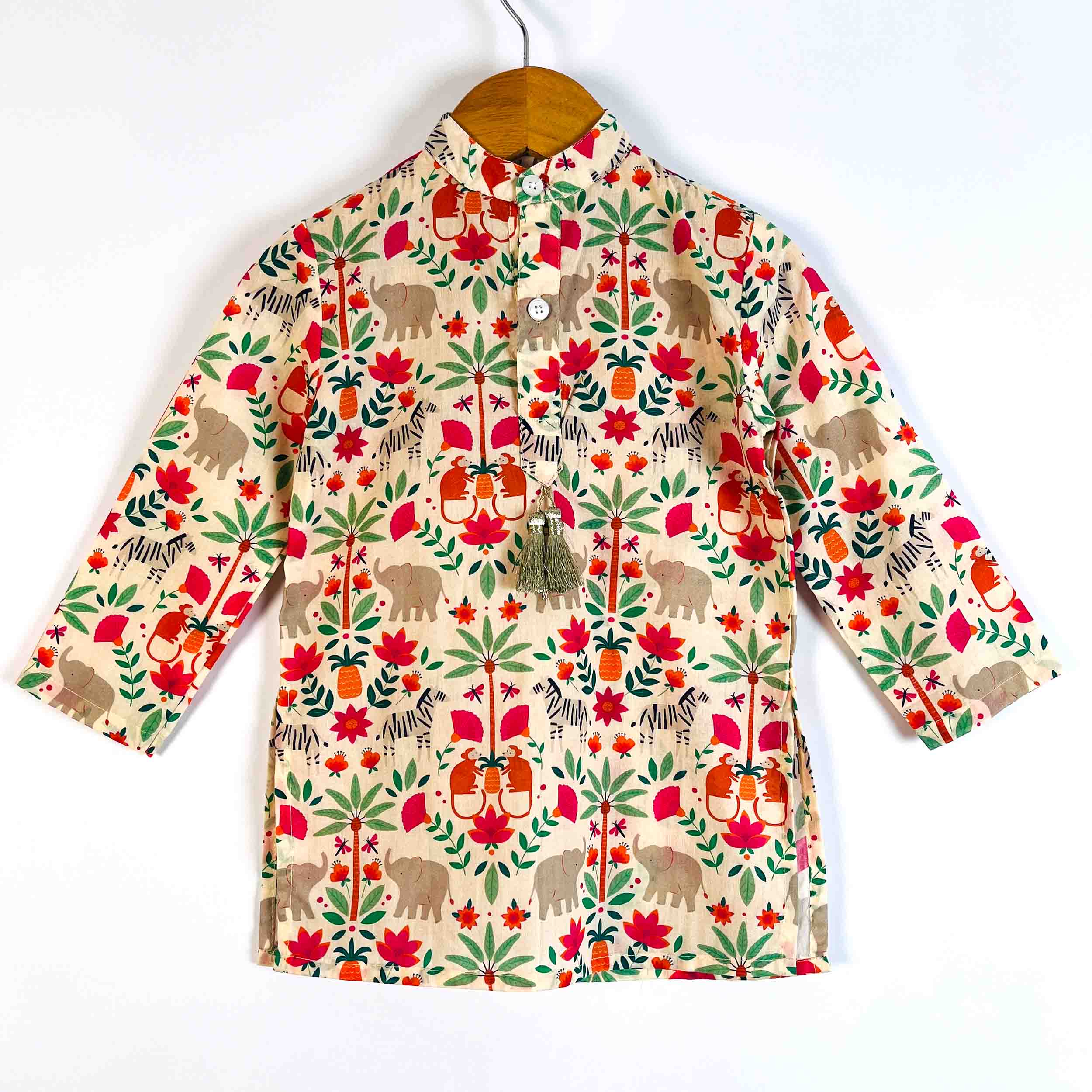 A long-sleeve button-up shirt for kids, featuring a colorful safari-themed print with red flowers, green leaves, zebra stripes, and cartoon elephants on a beige background, hanging on a wooden hanger against a white wall.