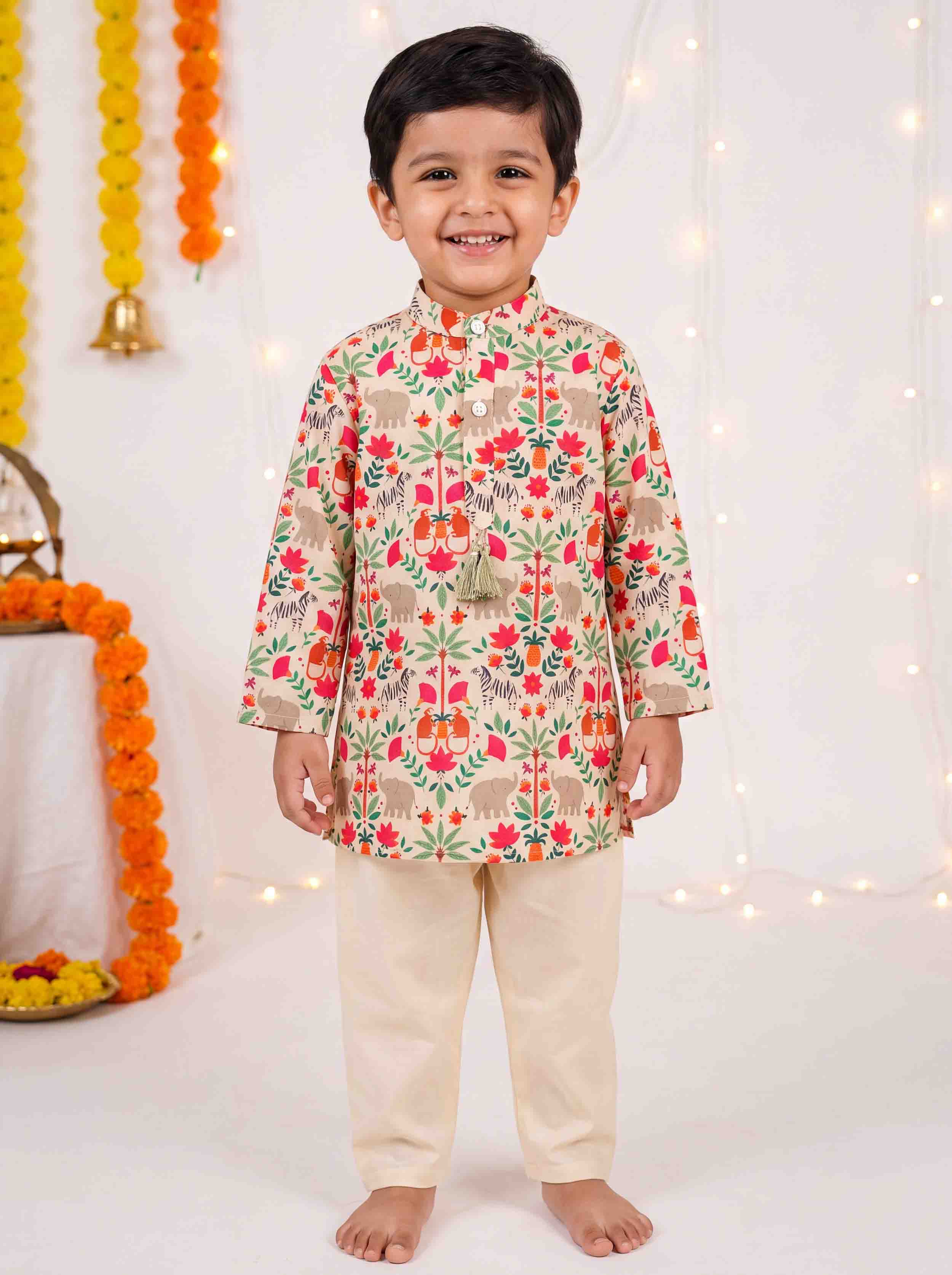 A young boy modeling a traditional Indian outfit consisting of a beige kurta with a colorful floral and elephant pattern, paired with solid beige trousers, set against a festive white backdrop with marigold garlands.