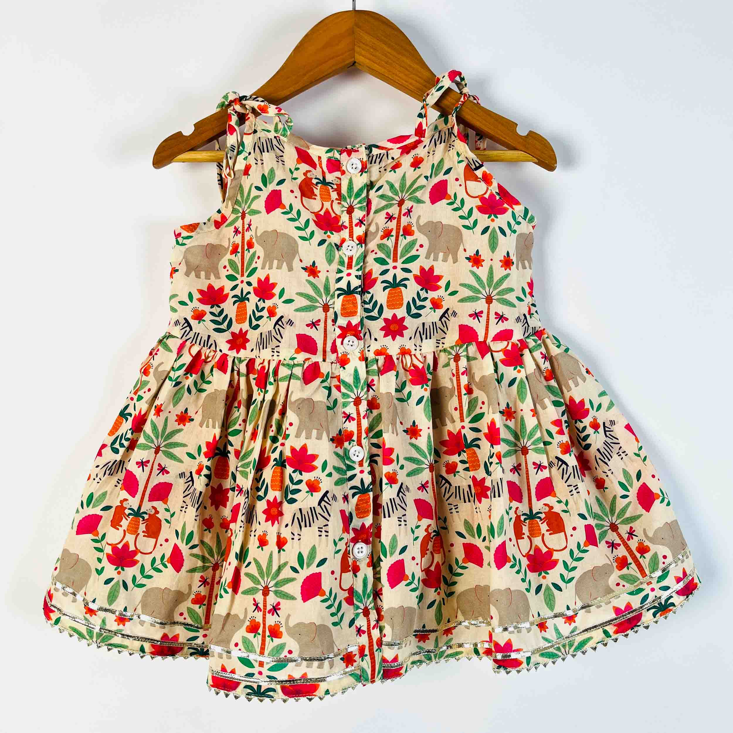 A sleeveless baby dress with a colorful safari print featuring elephants, zebras, and palm trees on a beige background, hanging on a wooden hanger against a plain white wall.
