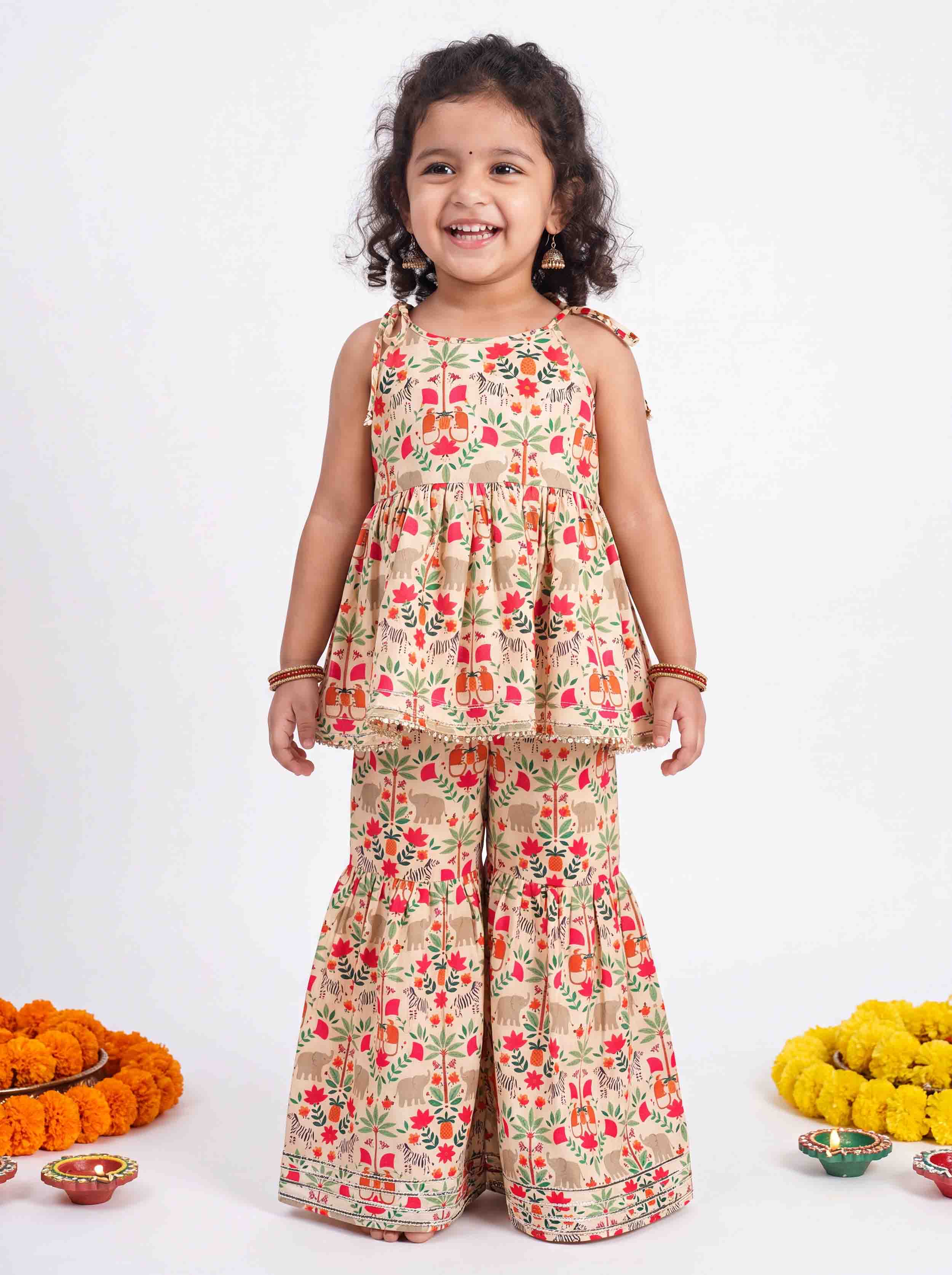 A young girl smiling and wearing a two-piece floral print outfit with a sleeveless peplum top and matching wide-leg pants. The outfit has a beige base with red and green floral patterns.
