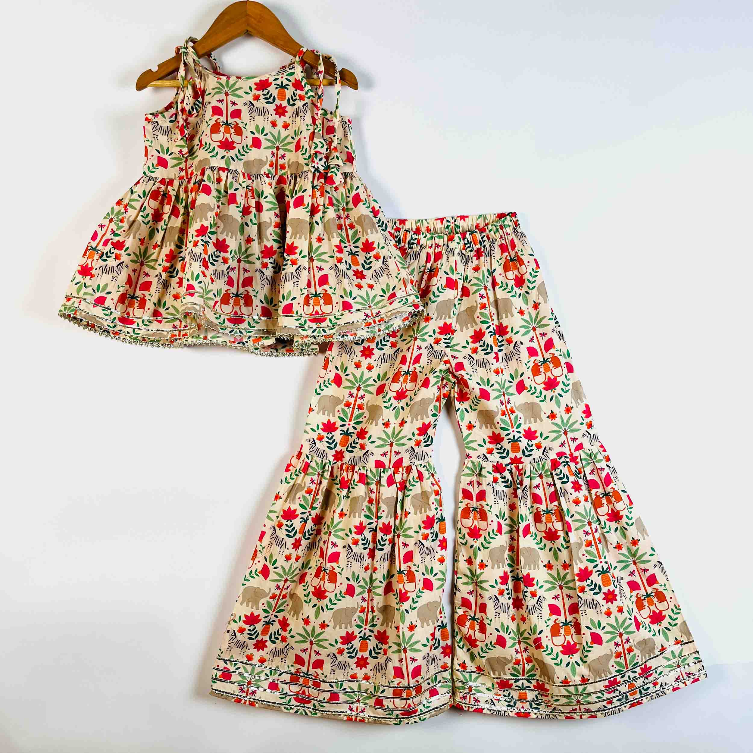 A two-piece children's outfit set consisting of a sleeveless ruffled top and matching wide-leg pants, both featuring a colorful floral and leaf print on a beige background, displayed on a wooden hanger and a flat surface against a white background.