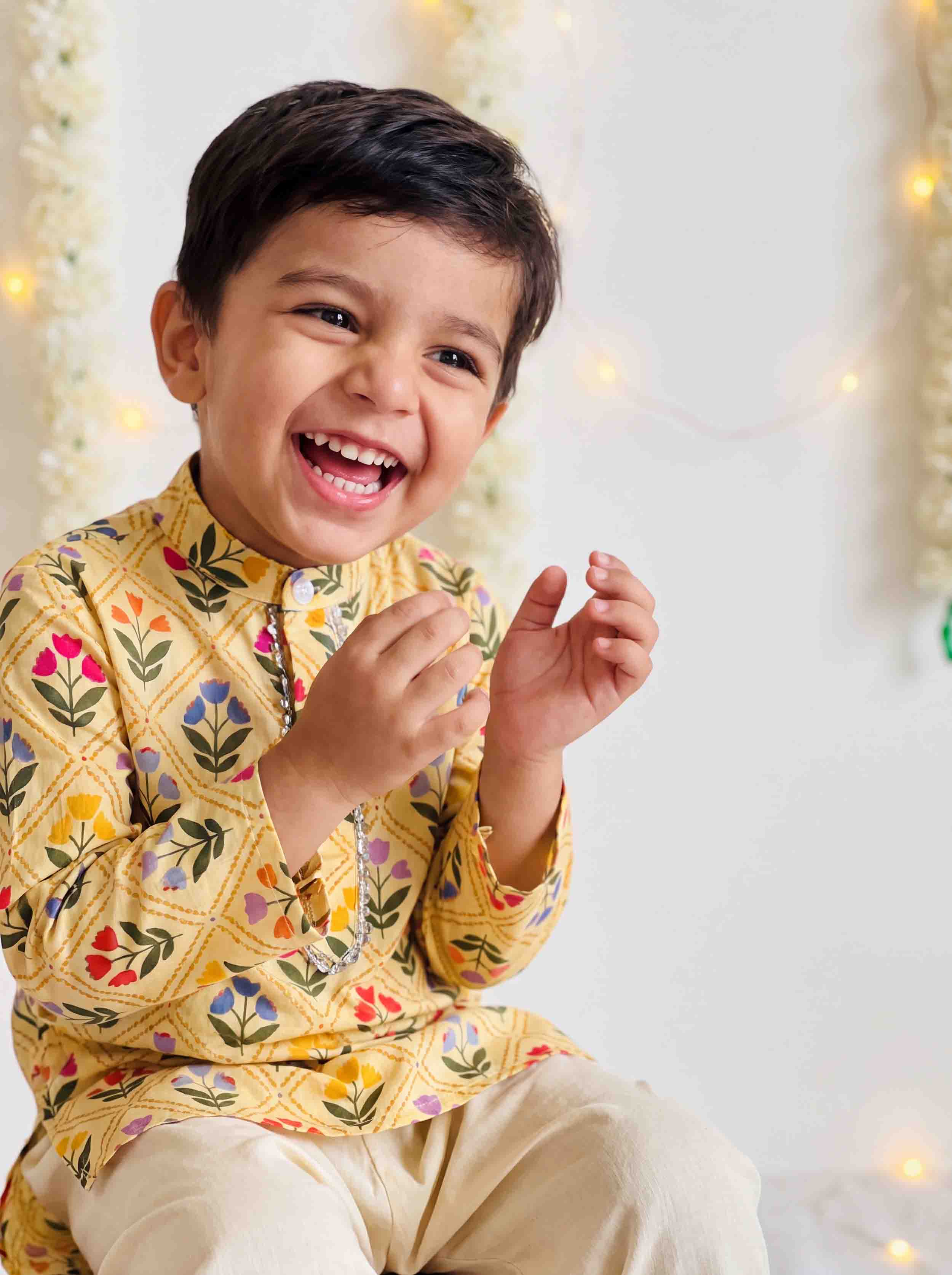 A young boy smiling and laughing while wearing a yellow embroidered kurta with colorful floral patterns and beige trousers, set against a softly lit white background with warm string lights.