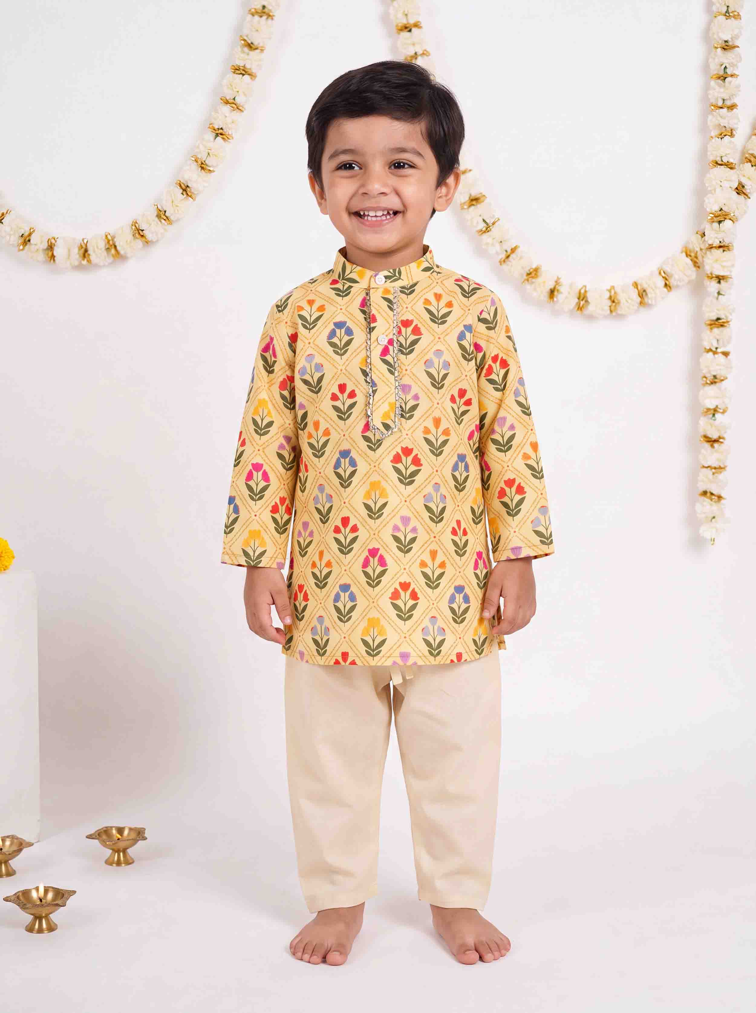 A young boy standing barefoot in a studio, wearing a yellow kurta with a colorful floral block print and beige trousers. The background is white with decorative garlands and small brass oil lamps on the floor.