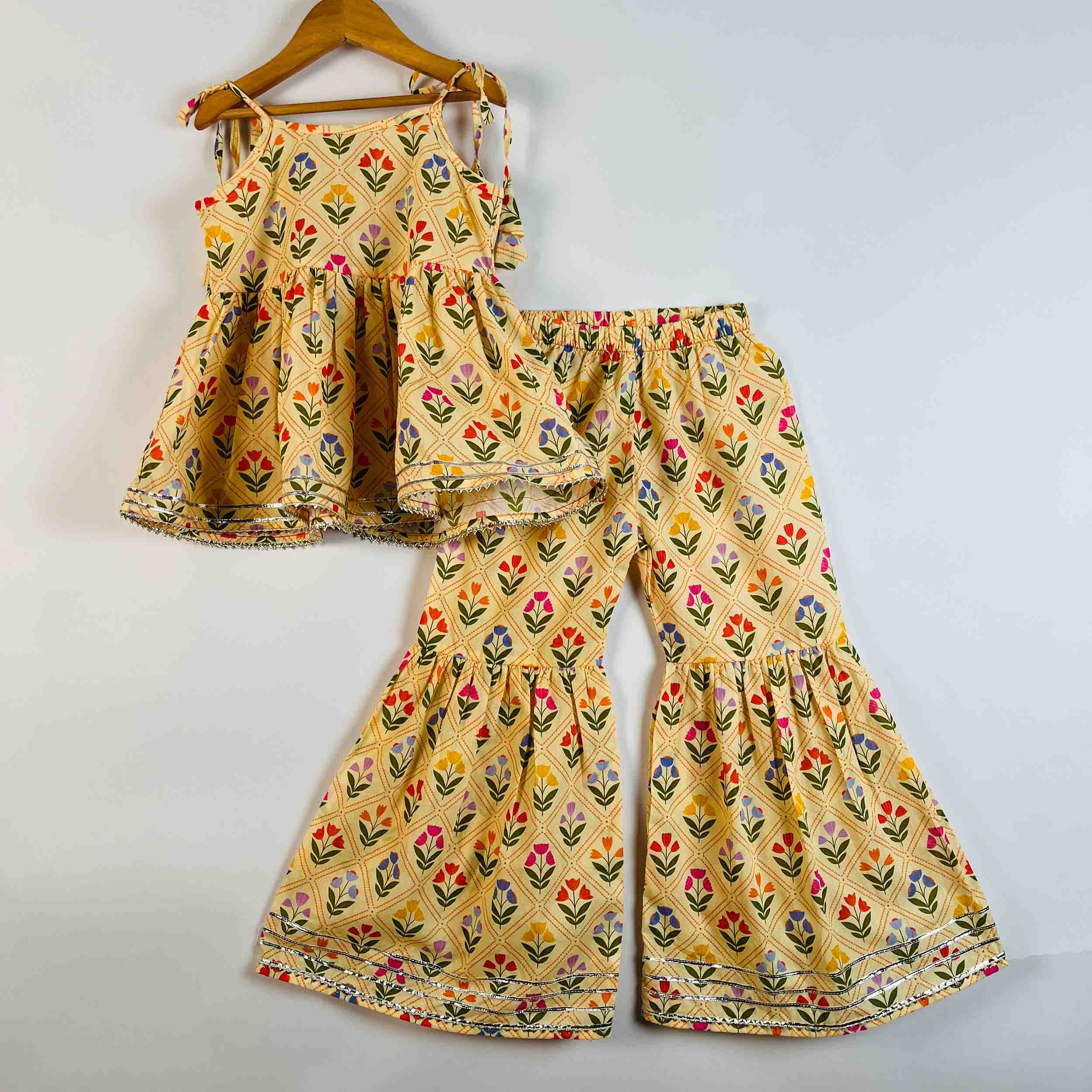 A two-piece children's outfit set featuring a sleeveless peplum top and matching wide-leg pants, both in a yellow fabric with a colorful floral block print, hanging on a wooden hanger against a plain light gray background.