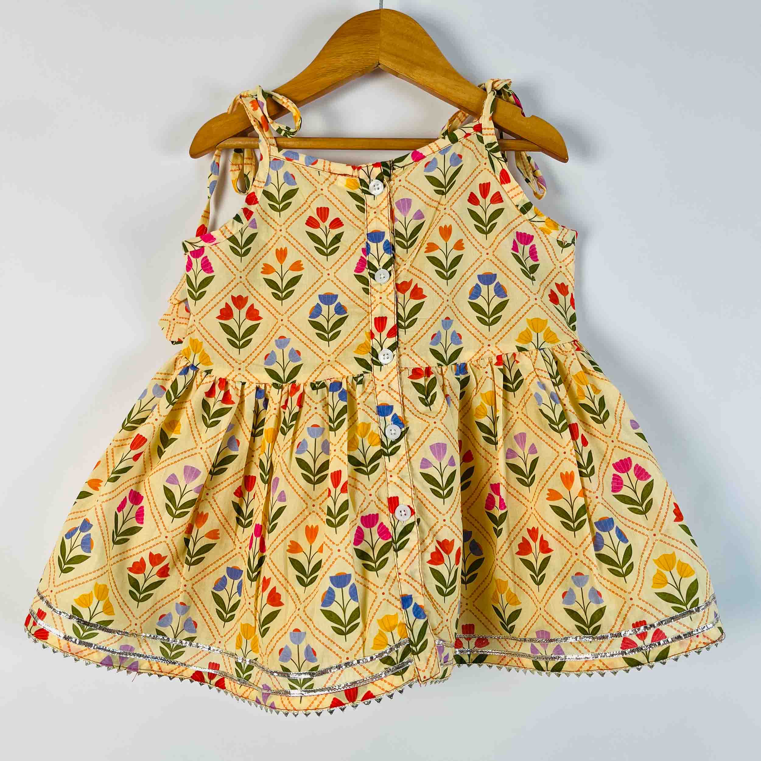 A toddler girl's sleeveless tiered dress with a colorful floral and diamond pattern on a cream-yellow background, hanging on a wooden hanger against a plain light gray wall.