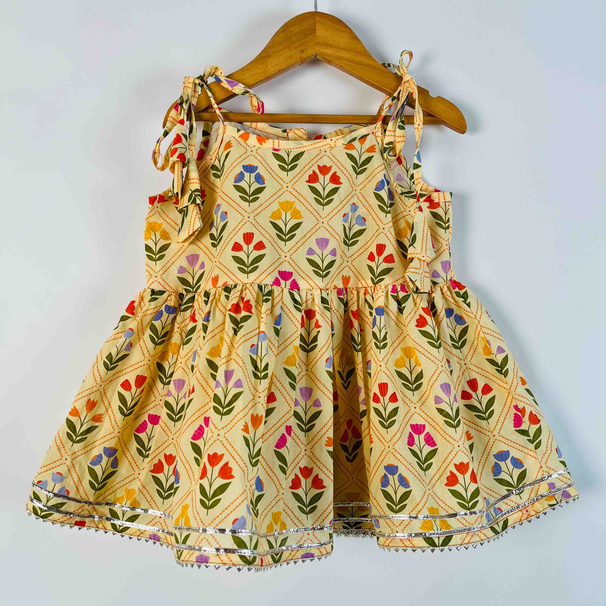 A sleeveless baby girl's dress with a cream-colored base and a colorful floral print, hanging on a wooden hanger against a light gray wall.