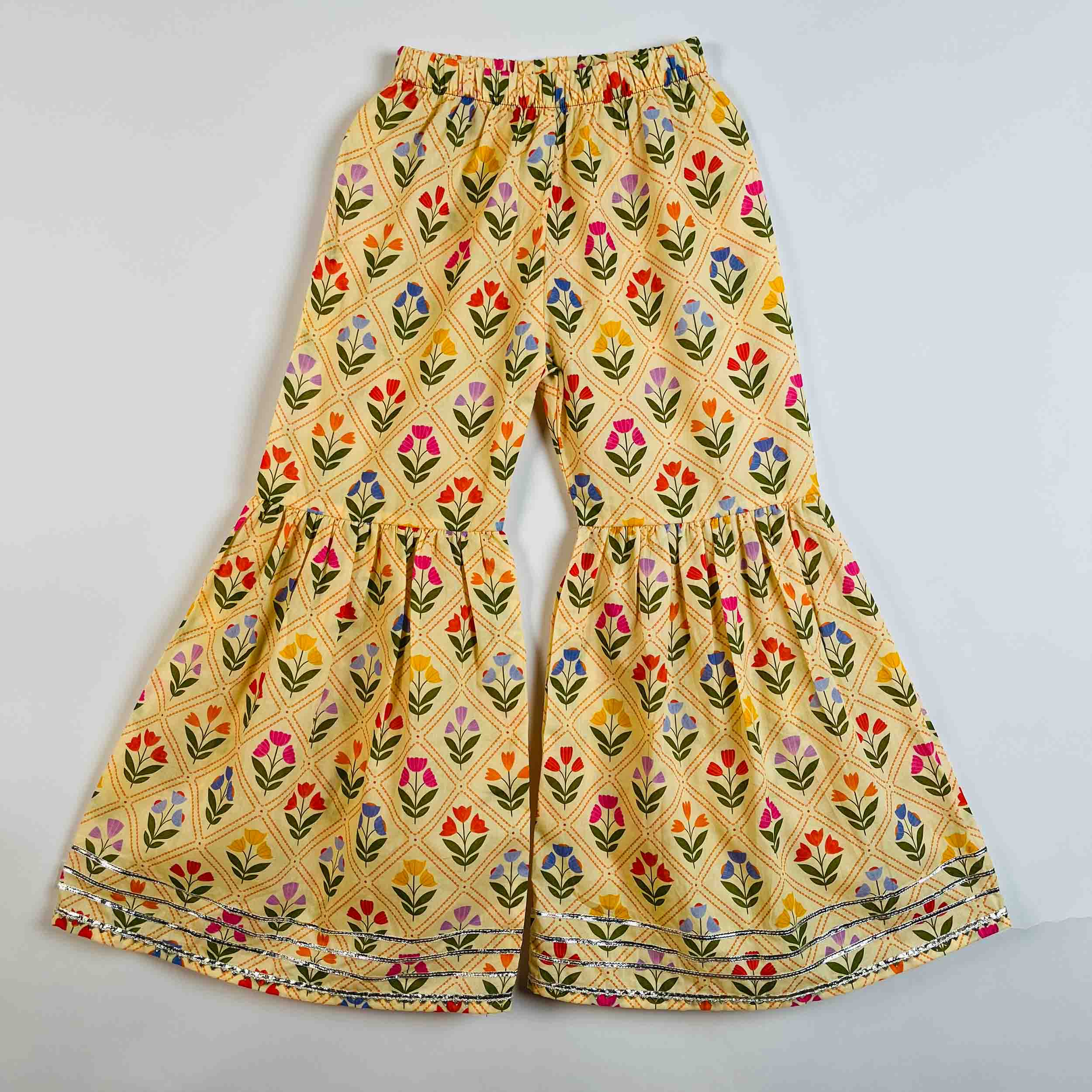 Yellow flared pants with a colorful floral block print pattern, featuring an elastic waistband and tiered ruffle legs, laid flat on a light gray background.