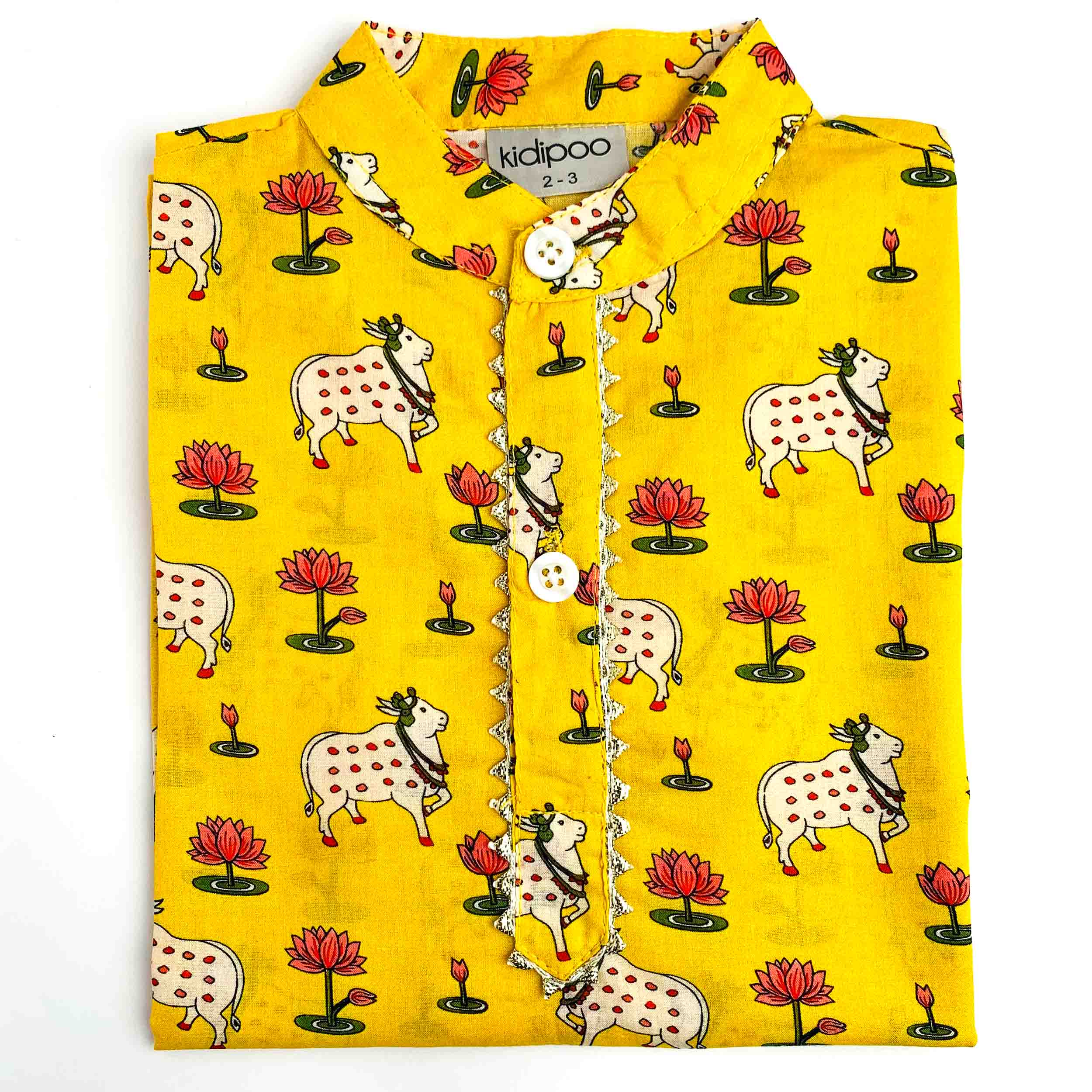 A folded yellow children's shirt with a printed pattern of cows, lotus flowers, and candles. The shirt features a mandarin collar, a button-up front, and a decorative silver chain detail down the center.